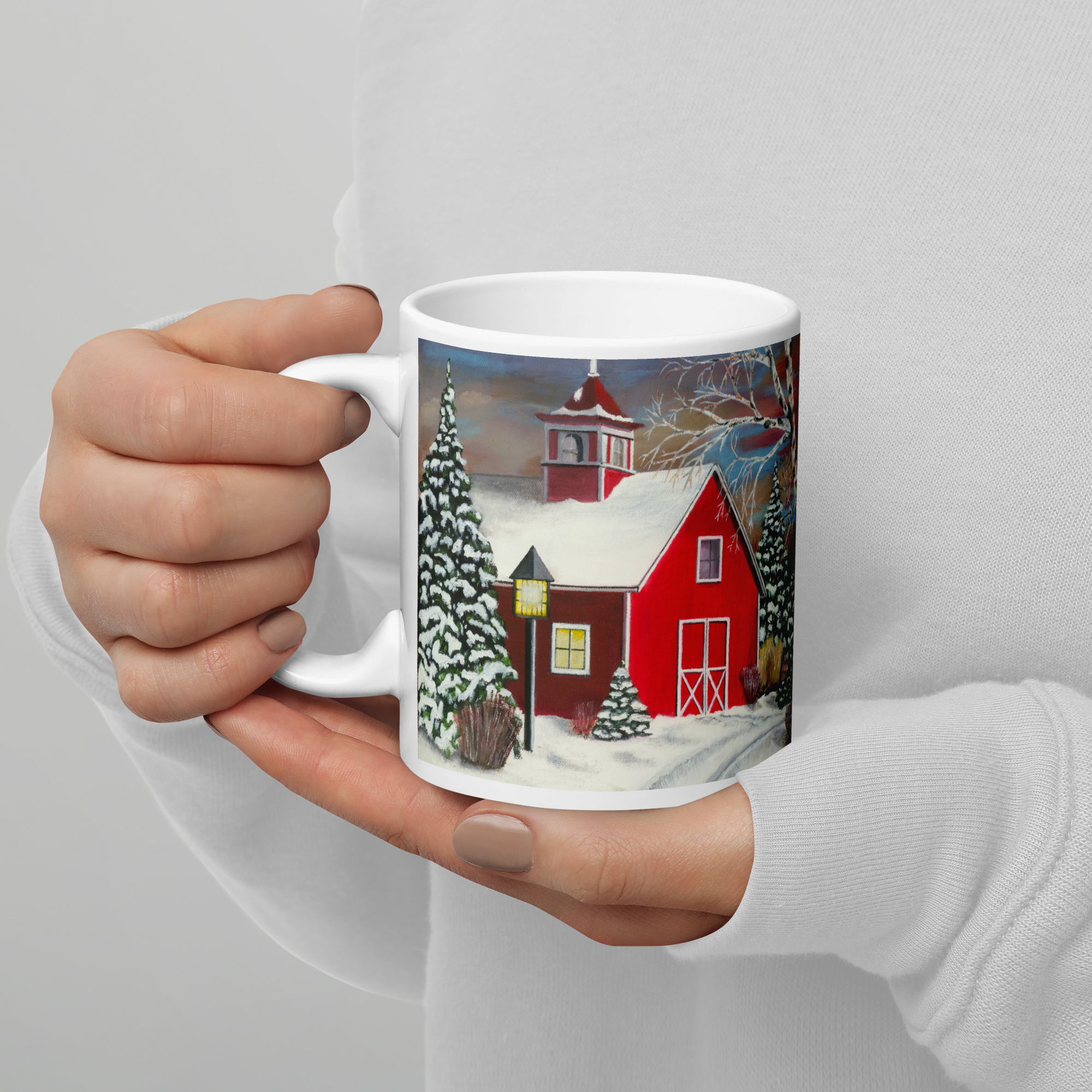 Art on a mug, Red Barn painting. A mug is held in two hands showing Landscape Artwork of a red barn in wintertime, covered in snow.